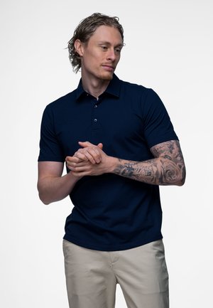 Man met tatoeages op zijn rechterarm, draagend een marineblauw poloshirt en beige broek, handen in elkaar geslagen en zijwaarts kijkend tegen een witte achtergrond.
