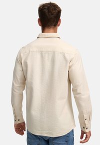 Chemise à manches longues de couleur crème avec une surface texturée, poignets boutonnés et col, associée à un jean bleu.