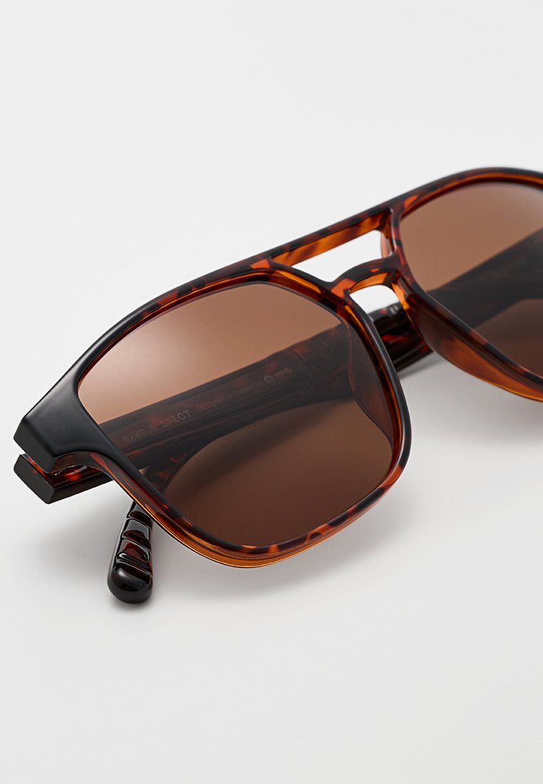 Słoneczne okulary w brązowym odcieniu z wzorem przypominającym skórę żółwia, mające grube, kanciaste oprawki i błyszczące czarne wykończenie na zausznikach.
