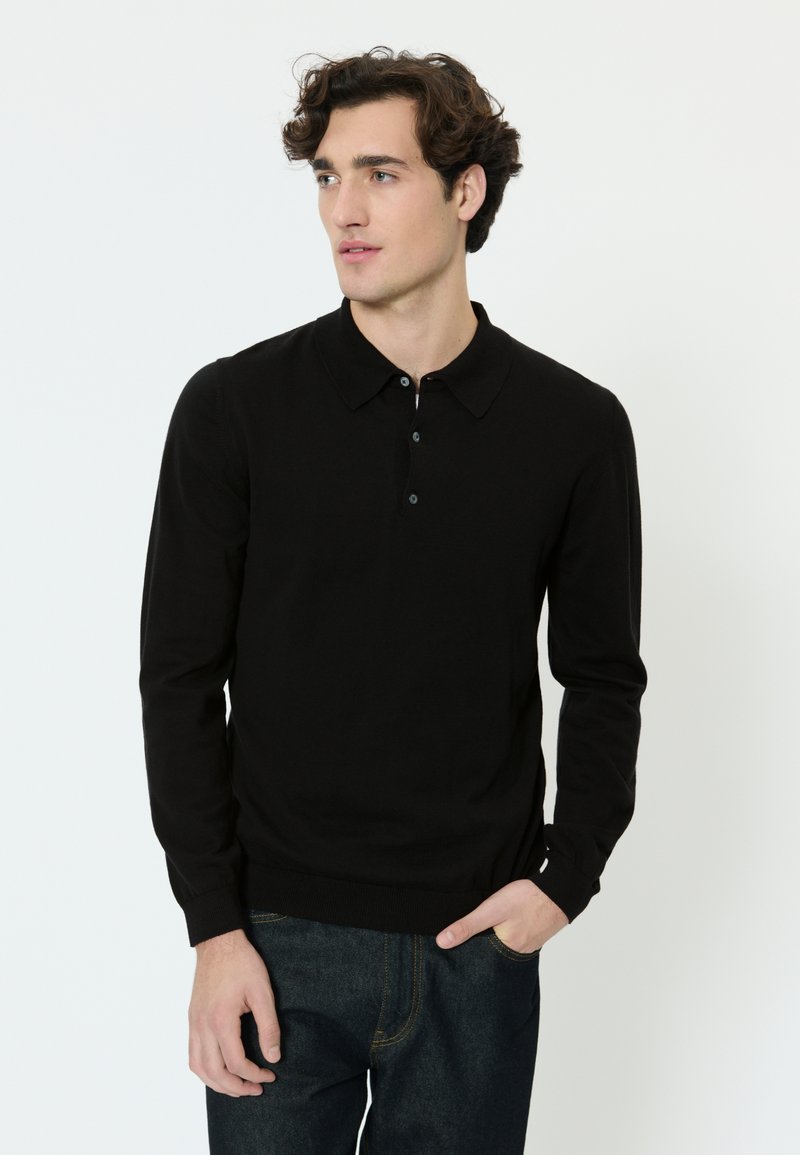 Muškarac s kovrčavom smeđom kosom odjeven u crnu polo majicu s dugim rukavima i tamne traperice, jedna ruka u džepu, gleda lijevo.