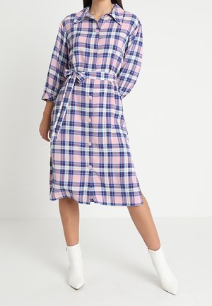 Woman wearing a pink and blue plaid button-down dress with a waist tie, paired with white ankle boots, standing with one leg bent.