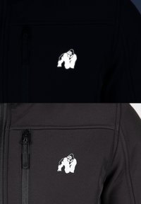 Veste softshell noire avec un logo de gorille réfléchissant blanc sur la poitrine. Comprend des poches zippées et un tissu lisse et texturé.