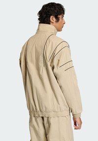 Μπεζ, ελαφρύ μπουφάν με ψηλό γιακά, ελαστική μέση και μαύρες ραφές κατά μήκος των ώμων και των μανικιών.