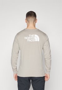 Graues Langarm-T-Shirt mit Rundhalsausschnitt, das ein großes weißes Logo im oberen Rückenbereich präsentiert. Glattes Material, lässige Passform.