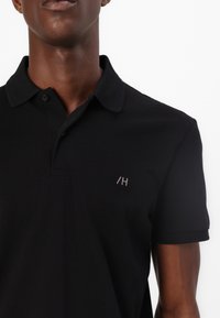 Camisa polo preta feita de tecido texturizado, com colarinho abotoado e um pequeno logotipo cinza claro no lado esquerdo do peito.