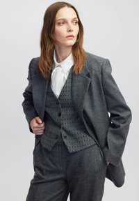 Grau gestrickter Blazer mit strukturierter Gestaltung, versehen mit einem taillierten Weste im Karomuster, getragen über einem weißen Hemd mit einer Krawatte.