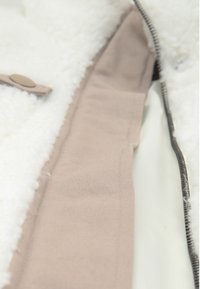 Hvid fleece-foring med en beige stofkant, udstyret med en lynlås og en brun knap. Blød tekstur med synlige sømme langs kanterne.