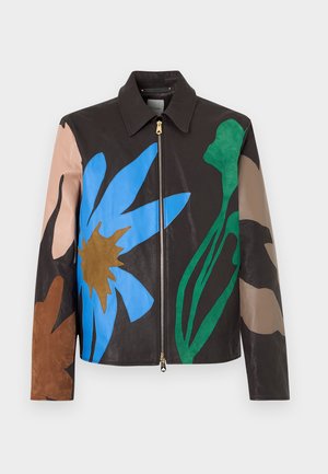 Jeune homme portant une veste en cuir noire avec de grands motifs floraux colorés, chemise grise, pantalon crème et baskets blanches, debout sur fond blanc.