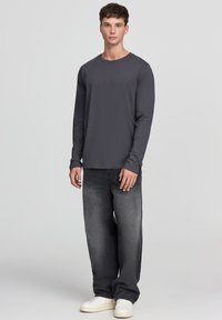 Maglietta grigia a maniche lunghe in cotone, abbinata a jeans in denim scuro e sneaker bianche. La vestibilità è rilassata e la maglietta ha un collo a giro.
