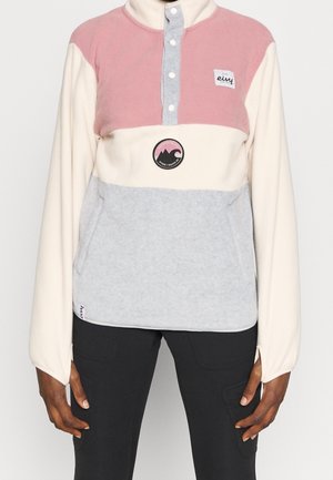 Persona que lleva un jersey polar con bloques de color en rosa, crema y gris, con botones y dos parches con el logo en el pecho.