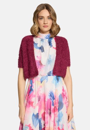 Femme aux cheveux blonds mi-longs portant une robe fleurie multicolore et un cardigan court texturé rose profond sur un fond uni.
