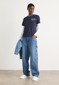 T-shirt navy con stampa "GANT 1948", sovrapposto a una maglietta a maniche lunghe bianca; abbinato a jeans larghi in denim chiaro e sneaker bianche.