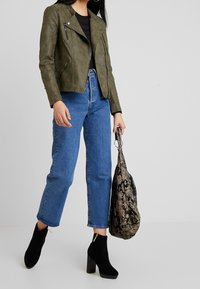 Frau trägt eine olivgrüne Zip-up-Jacke, hochtaillierte blaue Jeans, schwarze Stiefeletten mit Absatz und hält eine große Handtasche mit Schlangenledermuster.