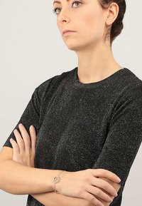 Top in maglia nera metallizzata con scollatura rotonda e maniche corte, indossato con un bracciale argentato che presenta un ciondolo circolare su una catena.