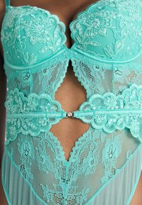 Turquoise lace bodysuit featuring floral embroidery, scalloped edges, mesh panels, and a shiny center clasp detailing.