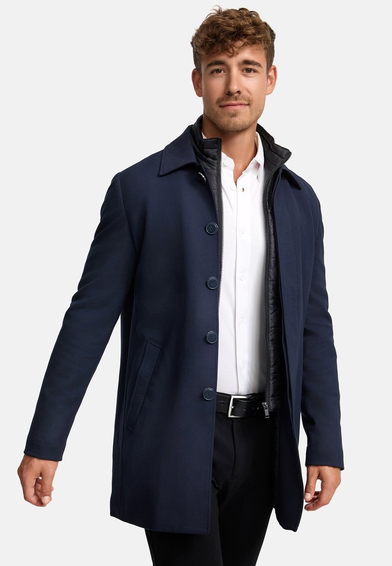 Jonge man gekleed in een marineblauwe jas met knopen, een zwarte binnenlaag met rits, een wit shirt, zwarte broek en een riem tegen een effen achtergrond.