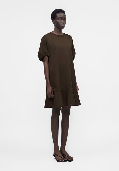 Γυναίκα που στέκεται φορώντας ένα φαρδύ, καφέ σκούρο φόρεμα με κοντά μανίκια και βολάν στο τελείωμα, καθώς και μαύρα σανδάλια με ανοιχτά δάχτυλα, μπροστά σε μια απλή ανοιχτή επιφάνεια.