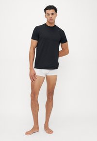 T-shirt noir à manches courtes texturé avec un col rond, associé à des sous-vêtements blancs, modèle se tenant avec une main sur la hanche, fond uni.