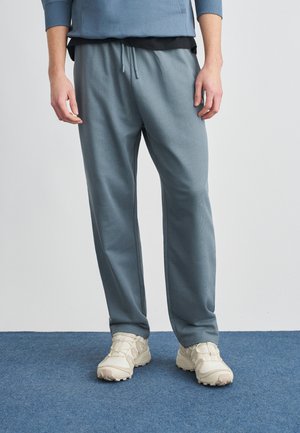 Uomo con pantaloni blu con coulisse, scarpe da ginnastica beige e una maglia blu a maniche lunghe, in piedi su un pavimento con moquette blu.