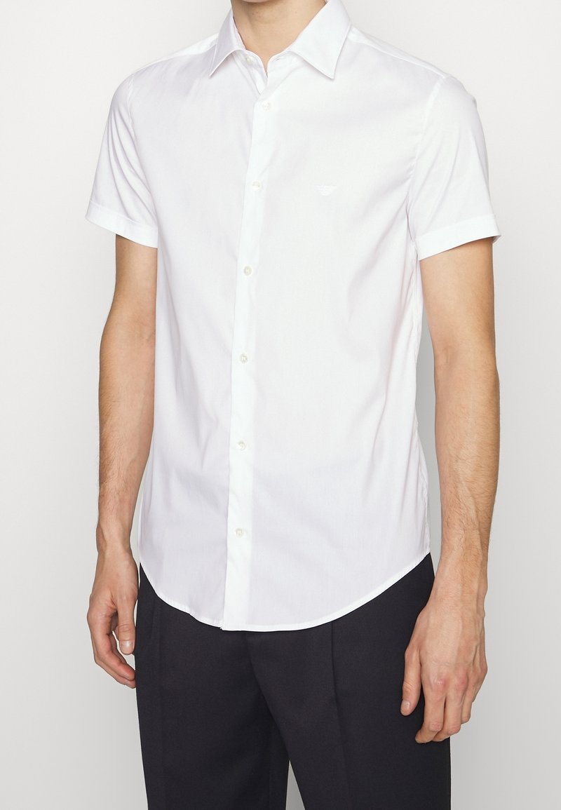 Muž v bílé košili s krátkým rukávem a zapínáním na knoflíky, oblečený v tmavých kalhotách, stojící na světlém jednobarevném pozadí.