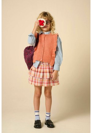 Enfant avec des lunettes tient un tissu avec des pompons rouges sur un œil, portant une chemise rayée, un gilet tricoté orange, une jupe à carreaux et des chaussures noires avec des chaussettes.
