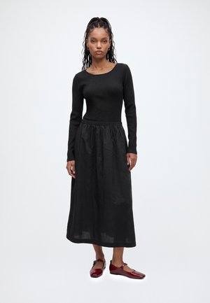 Femme debout portant un haut noir à manches longues, une jupe midi noire et des chaussures Mary Jane rouges sur fond blanc uni.