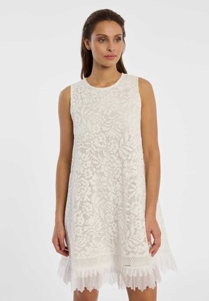 Vrouw draagt een mouwloze witte kanten jurk met bloemmotieven en een gerimpelde zoom, staand tegen een effen lichte achtergrond.