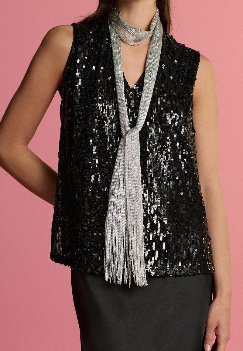 Femme portant un haut sans manches noir à sequins et un long collier argenté à franges sur fond rose.