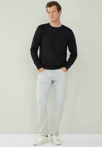 Sweatshirt en tricot noir avec un col rond, associé à un jean gris clair et des baskets blanches. Le modèle se tient de face avec les mains dans les poches.