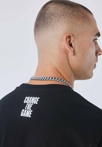 Camiseta negra con el texto "CAMBIA EL JUEGO" en blanco negrita en la parte superior de la espalda, complementada con un collar de cadena plateada.