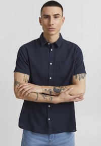Camisa de lino azul marino de manga corta con cuello abotonado, bolsillo frontal y textura suave. Modelo con tatuajes está de pie con los brazos cruzados.