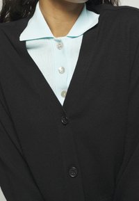Czarny kardigan z głębokim dekoltem w serek, z przodu zapinany na guziki i o prążkowanej fakturze, nałożony na jasnoniebieską koszulę z kołnierzykiem i detalami w postaci guzików.
