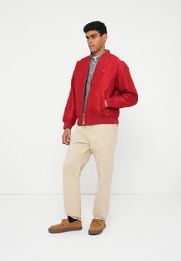 Punane pommitaja jakk ribidega varrukate ja hemiga, koos ruudulise särgi, beežide pükste ja pruunide sametjalatsitega.