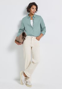 Donna che indossa una camicia verde menta con bottoni, una canotta bianca, pantaloni larghi color crema e sneaker bianche, mentre tiene una borsa beige contro uno sfondo bianco.