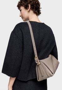 Mujer con cabello corto y rizado, vestida con un suéter oscuro de gran tamaño y llevando un bolso de hombro de cuero taupe con hebilla dorada.