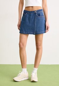 Μίνι σορτσάκι από τζιν σε μπλε χρώμα με εφαρμοστή μέση, δύο μπροστινές τσέπες και αντίθετη ραφή. Συνδυασμένο με ανοιχτό μπέζ αθλητικά παπούτσια και λευκές κάλτσες.