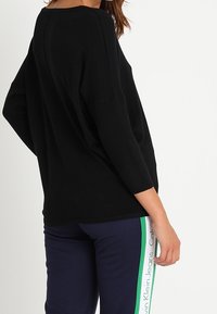 Svart oversized tröja med ribbad struktur och trekvartsärmar, tillsammans med mörka byxor med gröna och vita sidostripes.