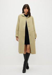 Cappotto di lana beige con colletto arrotondato, cinque bottoni, due tasche frontali e maniche arrotolate, abbinato a un vestito nero lavorato a maglia e stivali neri al ginocchio.