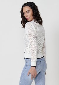 Suéter blanco de punto con patrón de encaje geométrico, puños acanalados y dobladillo. Llevado sobre unos pantalones vaqueros de un azul claro. El modelo posa de lado, con el cabello suelto.