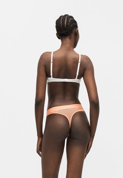 Γυναίκα με πλεξούδες στα μαλλιά, φοράει λευκό στηθόδεσμο και ροδακινί ριμπεζέ κορδόνι, στέκεται μπροστά σε ένα απλό ανοιχτό γκρι φόντο, από πίσω.