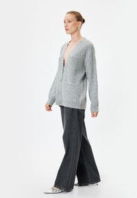 Ljusgrå stickad cardigan med djup V-hals, framfickor och knäppning, kombinerad med vida mörka denimjeans och metalliska skor.