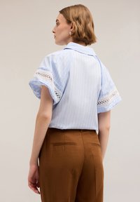 Jasnoniebieska koszula w paski z koronkowym wykończeniem na krótkich rękawach, w połączeniu z brązowymi spodniami z wysokim stanem. Gładki materiał, klasyczny krój kołnierzyka.
