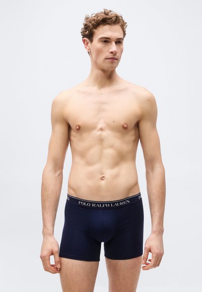 Άνδρας χωρίς πουκάμισο με κατσαρά μαλλιά που φοράει σκούρα μπλε boxer briefs Polo Ralph Lauren, στέκεται μπροστά σε απλό ανοιχτόχρωμο φόντο.