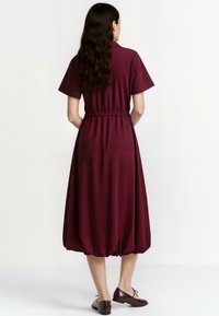 Burgundin mekko, jossa on lyhyet hihat ja joustava vyötärö, ja joka sisältää ilmavan alaosan. Yhdistetty ruskeisiin tasapohjaisiin kenkiin. Sujuva kankaan texture.