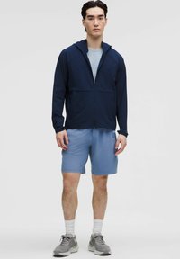 Marineblå zip-up træningsjakke med hætte, parret med lyseblå shorts. Modellen har grå sneakers og hvide sokker på, hvilket fremhæver en strømlinet pasform.