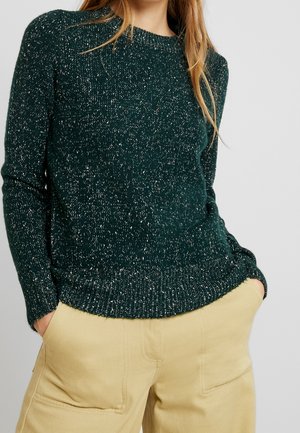 Vrouw die een donkergroene gebreide trui met spikkels en beige broek draagt, met haar handen in de zakken, tegen een effen lichte achtergrond.