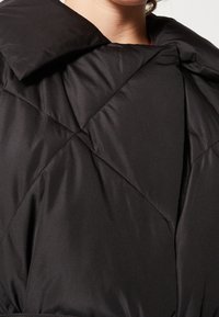 Detailní záběr na límec a oblast hrudi černé prošívané bundy na osobě s viditelným částečným krkem.