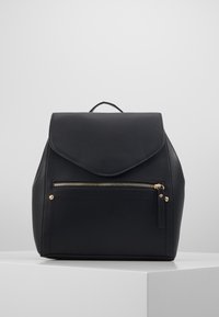 Sac à dos en cuir noir avec une fermeture à rabat, une poche zippée à l'avant, des accents en métal doré et une poignée supérieure pour le transport.