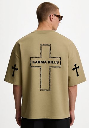Homme portant un t-shirt oversize kaki avec des croix noires sur les manches et le texte "KARMA KILLS" formant une grande croix dans le dos.
