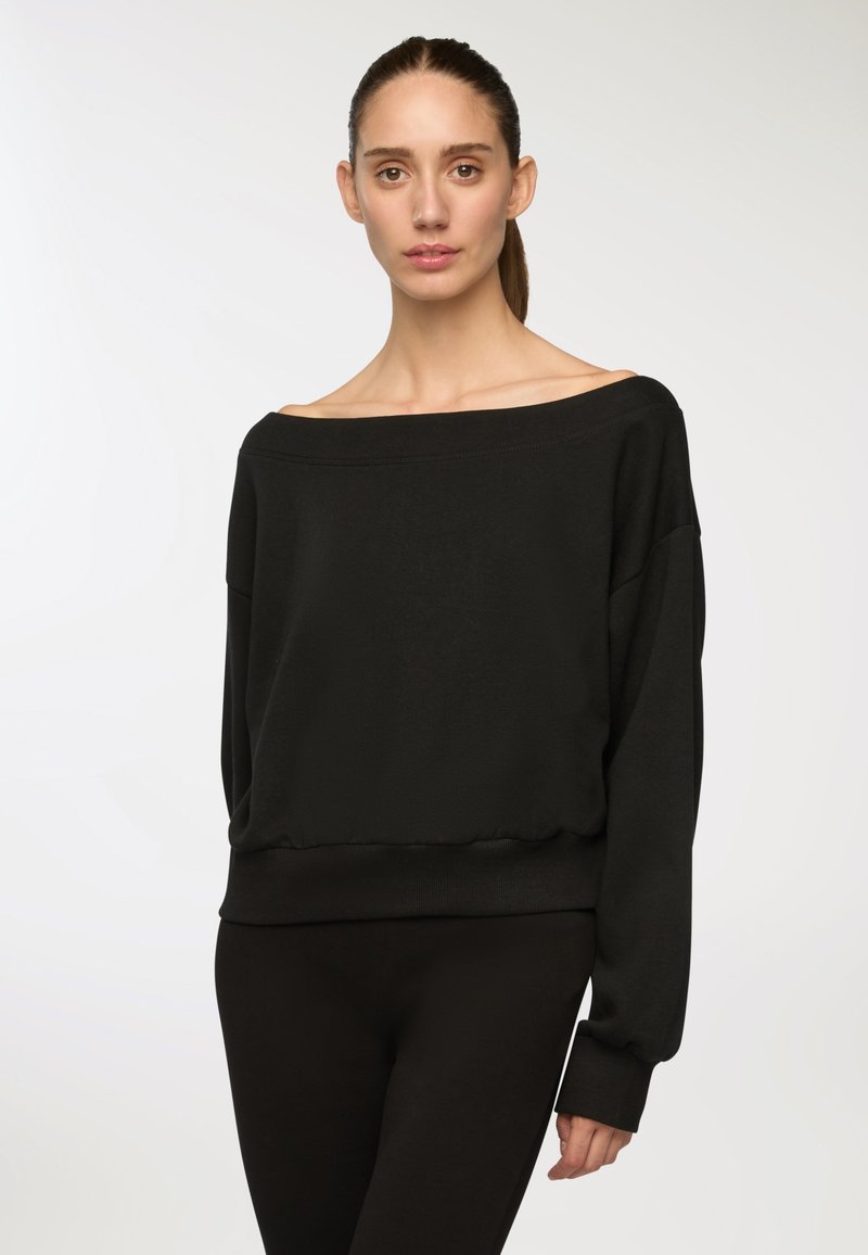 Felpa nera off-the-shoulder con vestibilità relaxed, polsini e orlo a coste. Realizzata in tessuto morbido con una texture liscia, abbinata a leggings neri.